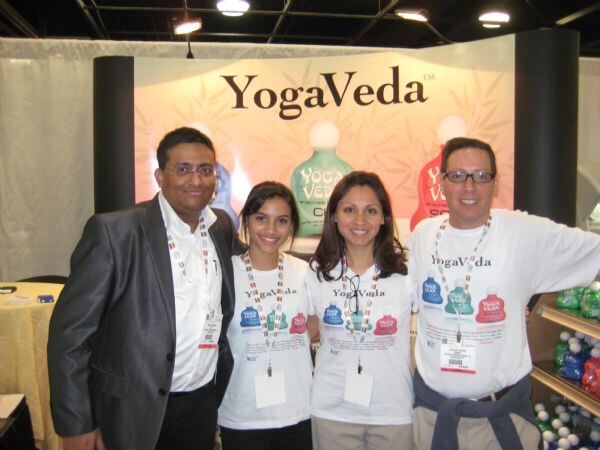 Four people, a man in a gray blazer, two women and a man in glasses, stand together in front of a YogaVeda sign; the women and one man wear YogaVeda t-shirts.