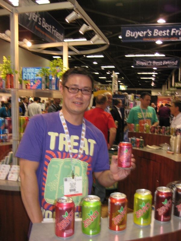 A man in glasses and a purple Treat t-shirt holds a can of Zevia soda behind a counter lined with colorful Zevia cans, with other attendees and booths behind him.