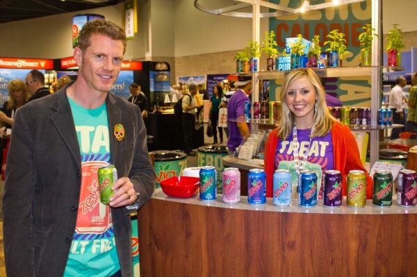 A man in a teal Zevia shirt and gray blazer holds a can, and a smiling blonde woman in an orange cardigan stands beside a counter lined with Zevia soda cans.
