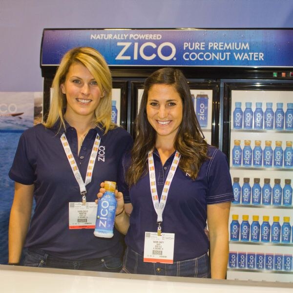 Two smiling women in navy ZICO polo shirts and lanyards stand at a counter; one holds a bottle of ZICO coconut water in front of refrigerated cases and a ZICO sign.