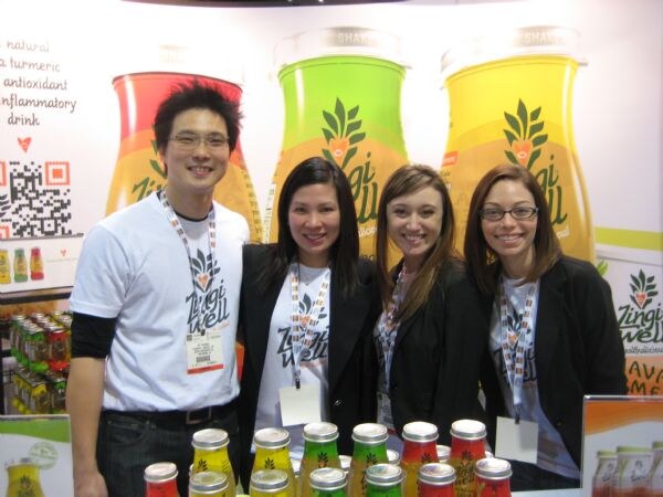 Four smiling people wearing lanyards, a man in a white Zingi Well t-shirt and three women, stand behind small bottles in front of a Zingi Well banner.