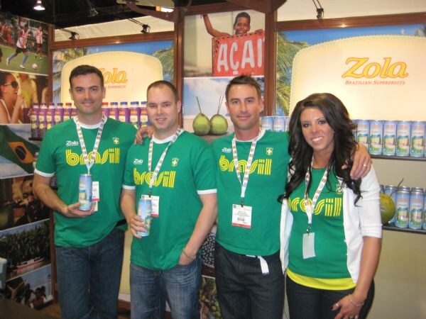 Three men and a woman in green brasil t-shirts and lanyards pose at the Zola booth; some hold cans, and shelves of Zola products and an açaí sign are behind them.