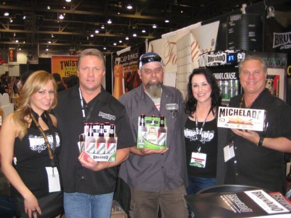 Five people with lanyards stand together at a trade-show booth; two men hold six-packs of Bruvado beer and another holds a Michelada box, with a Shoot Chase sign behind.