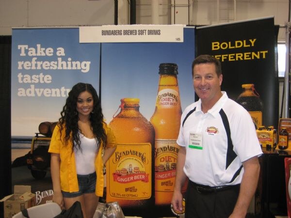 A smiling woman in a yellow jacket and a smiling man in a white and black polo stand in front of Bundaberg Brewed Soft Drinks banners showing Ginger Beer and Diet Ginger Beer bottles.