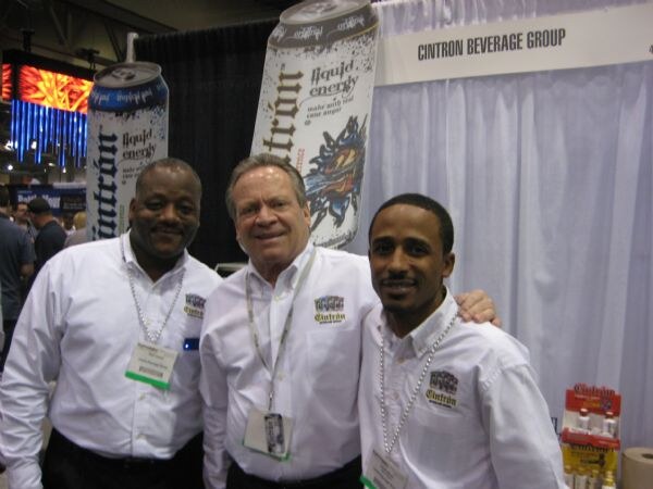 Three men in white Cintron shirts and lanyards pose together under large Cintron liquid energy can displays, with a Cintron Beverage Group sign above.