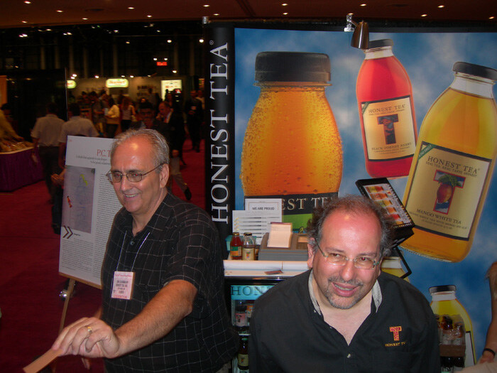 Two men with glasses stand at an Honest Tea booth in front of large banners of bottled teas. The man at right wears a black Honest Tea shirt. Both wear badges.