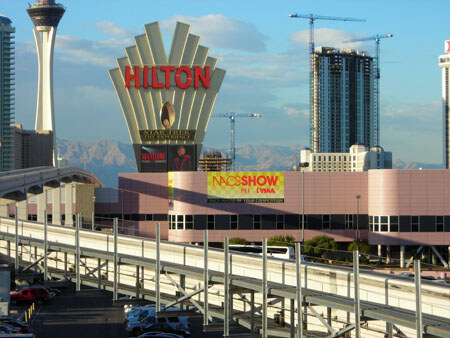 Tall Hilton sign with red letters in front of a pink building with a NACS Show banner, an elevated track in the foreground, construction cranes, a tower and mountains behind.