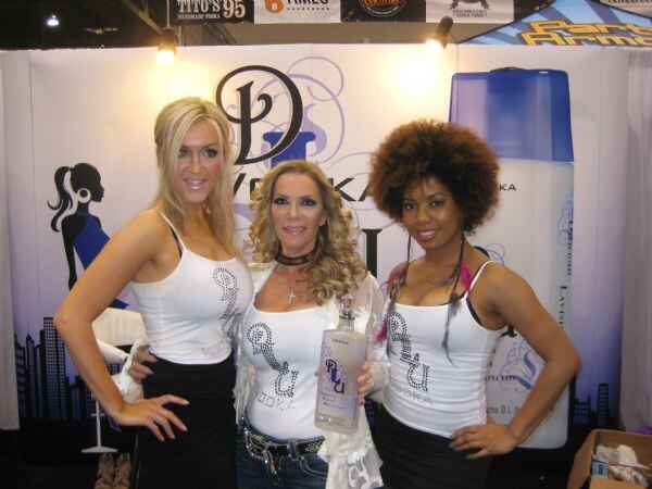 Three women in white DLU Vodka tank tops pose at the booth, the middle one holding a bottle of DLU Vodka, with a Tito's Handmade Vodka sign visible above.