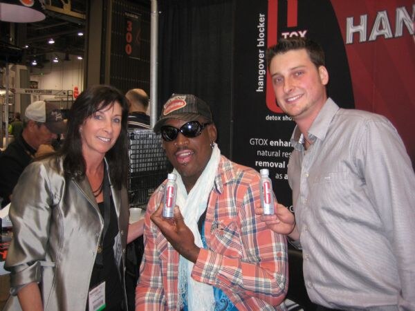 A woman, a man in sunglasses and a plaid shirt, and a second man pose at the GTOX booth; the two men hold small GTOX bottles in front of a hangover blocker sign.