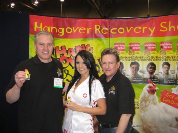 Two men in black Hangover Joe's shirts and a woman in a white nurse-style outfit pose at the booth, with small shot bottles in hand, before a Hangover Recovery Shot banner.