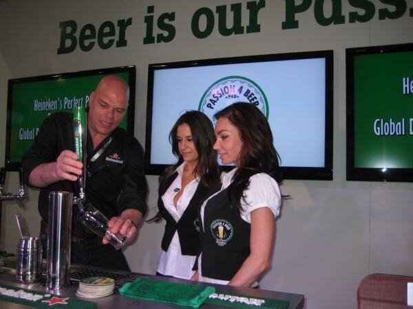 A bald man in a black Heineken shirt pours a beer from a tap into a glass while two women in Passion 4 Beer uniforms watch, with screens showing Heineken and Passion 4 Beer branding.