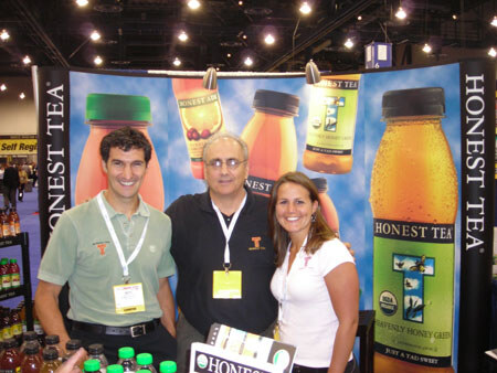 Two men and a woman with lanyards pose together in front of an Honest Tea banner showing bottles of tea, with bottles of Honest Tea on a shelf at left.