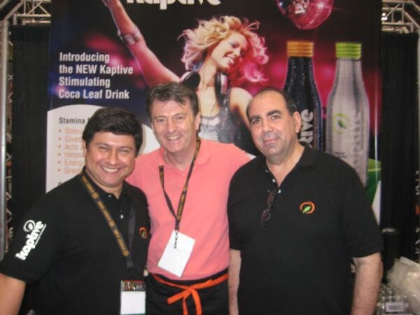 Three smiling men pose in front of a Kaptive banner for a stimulating coca leaf drink. Two wear black Kaptive polo shirts and one wears a pink shirt with a lanyard and name badge.