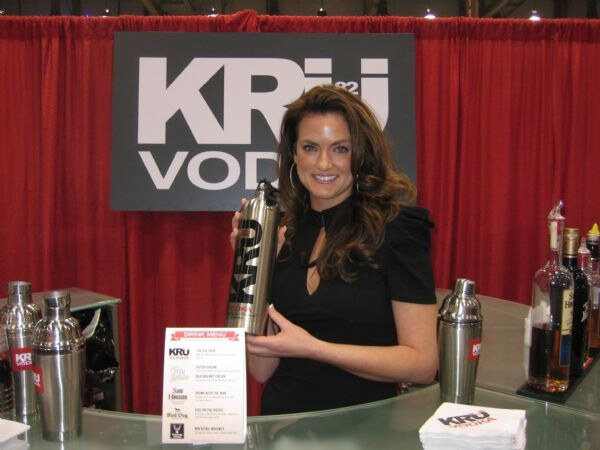 A smiling woman with long brown hair in a black top holds a tall steel KRU Vodka bottle behind a glass bar. A KRU Vodka sign, cocktail shakers and a drink menu listing KRU Vodka, Sam Hanson and Blind Dog are visible.