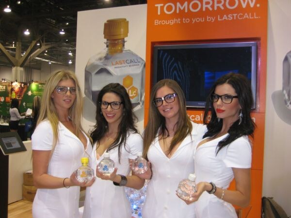 Four women in white outfits and dark-rimmed glasses hold small water bottles in front of an orange LastCall display with a large bottle graphic and a screen reading Tomorrow.