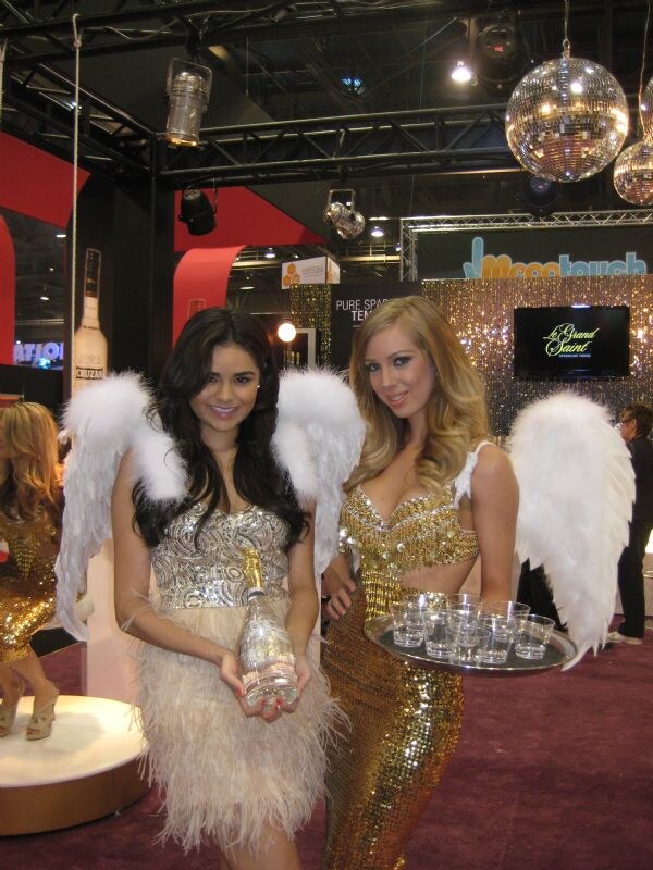 Two women in feathered white angel wings pose in a booth. One in a sequined dress holds a bottle, the other in a gold sequined outfit holds a tray of small cups. Le Grand Saint and Mood Touch signs are behind them.