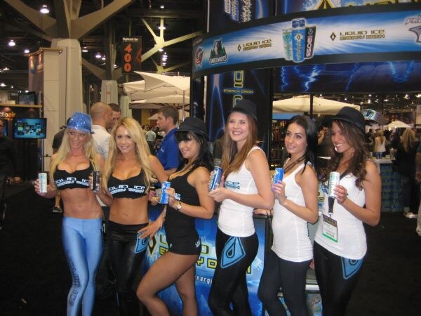 Six women in Liquid Ice branded tops and leggings stand in a row holding cans of Liquid Ice energy drink in front of the blue Liquid Ice booth, with attendees visible behind them.