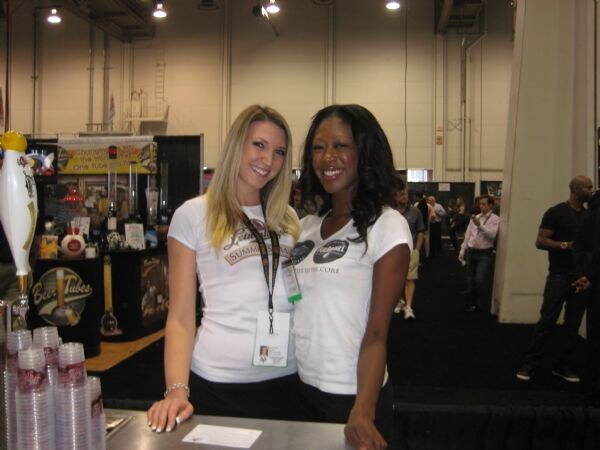 Two smiling women in white t-shirts stand behind a counter at the Beer Tubes booth. Stacks of plastic cups are at left, and a tap-style display and attendees are in the background.