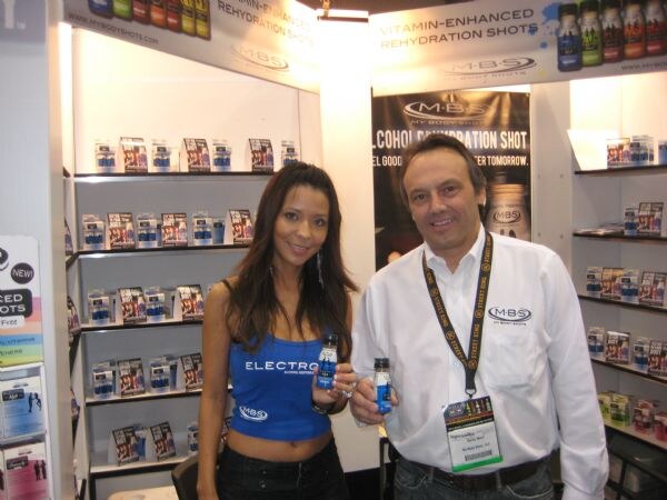 A woman in a blue M·B·S top and a man in a white shirt with a lanyard each hold a small bottle in front of shelves of M·B·S My Body Shots rehydration shot packages.