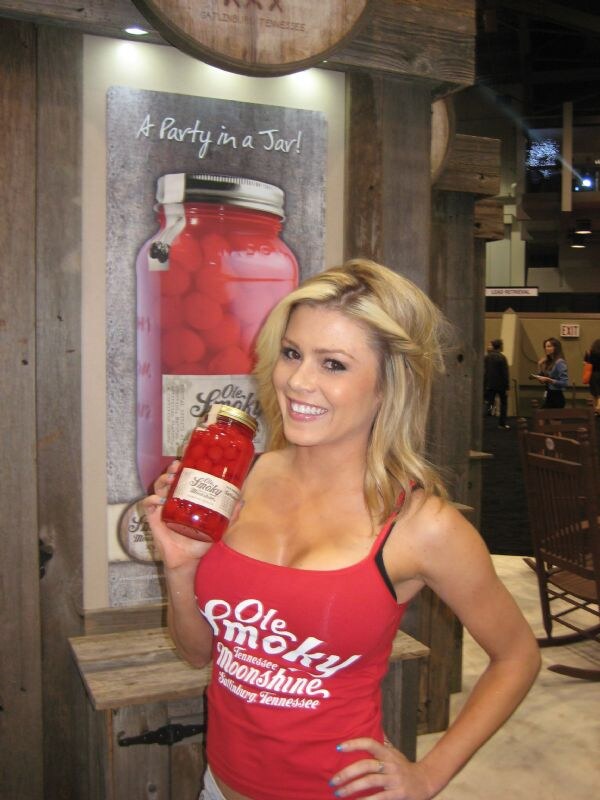 A smiling blonde woman in a red Ole Smoky Tennessee Moonshine tank top holds a jar of cherries in moonshine in front of a wooden booth with a poster reading A Party in a Jar!