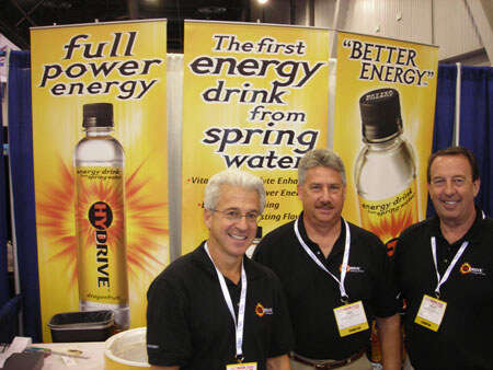 Three men in black polo shirts and lanyards stand in front of yellow HyDrive banners reading 'Full power energy' and 'The first energy drink from spring water'.