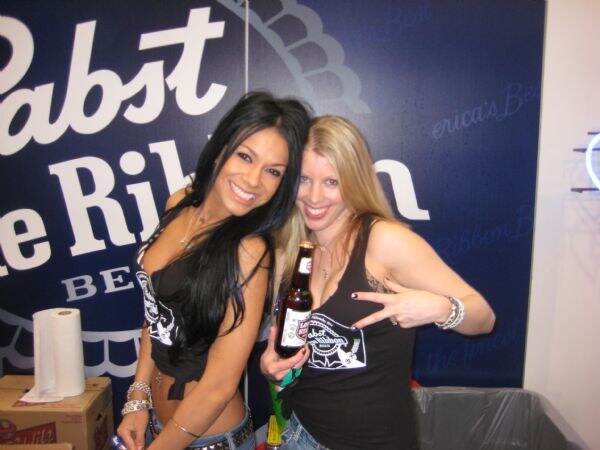 Two smiling women in black Pabst Blue Ribbon tank tops pose in front of a blue Pabst banner; one holds a bottle of beer and the other makes a hand gesture.