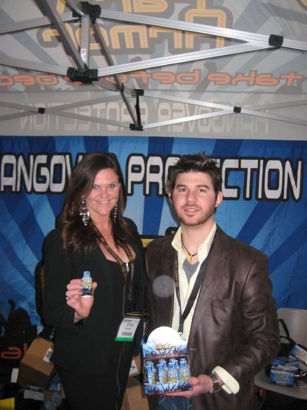 A woman and a man in a tent booth hold a small bottle and a display box of Party Armor shots in front of a blue Hangover Protection banner.