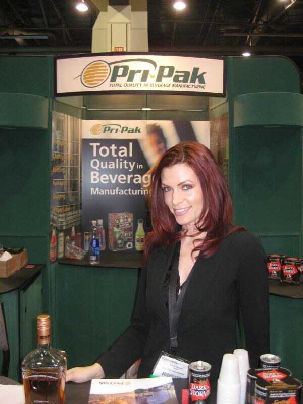 A red-haired woman in a black blazer stands in the green Pri-Pak booth; Gosling's Dark 'n Stormy cans, a whiskey bottle and a brochure sit on the counter.