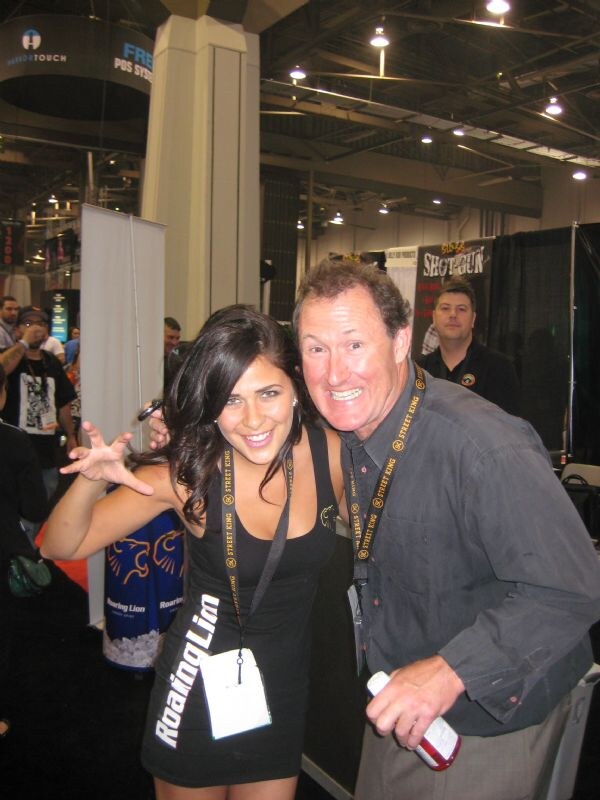 A woman in a Roaring Lion dress makes a clawing gesture beside a smiling man; both wear Street King lanyards on a trade show floor with a Roaring Lion display behind.