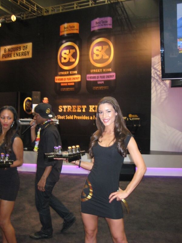A woman in a black dress holds a tray of shot bottles in front of Street King 6 Hours of Pure Energy displays; other staff walk behind her.