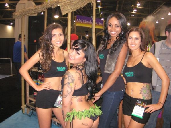 Four women in black Ganja Girls tops pose together under a thatched roof booth structure; one wears a green grass-style skirt.