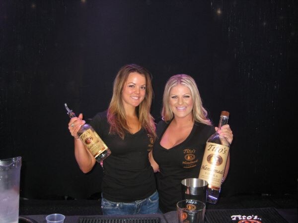 Two smiling women in black Tito's Handmade Vodka shirts stand behind a bar, each holding up a bottle of Tito's Handmade Vodka.