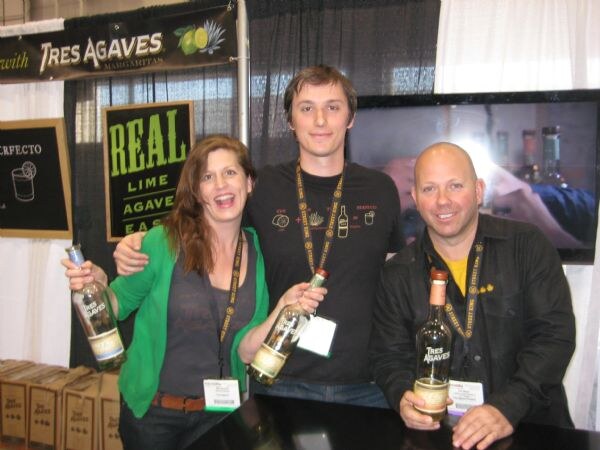 Two men and a woman behind a counter hold bottles of Tres Agaves tequila, with a Tres Agaves banner and a Real Lime Agave sign behind them.