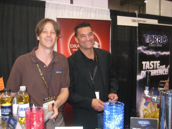 Two men stand at the Turbo Energy Drink booth with bottles and cups on the counter, a tap on the right and a Turbo banner reading Taste the Difference.