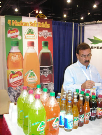 A man with a mustache looks down at a table lined with colorful Jarritos, Mundet and Señorial soda bottles beneath a Mexican soft drinks banner.
