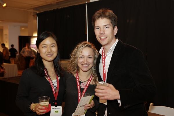 Three smiling attendees pose together at an indoor event, wearing red lanyards. The woman on the left, Amy, holds a glass of beer; the woman in the middle and a man on the right hold drinks.