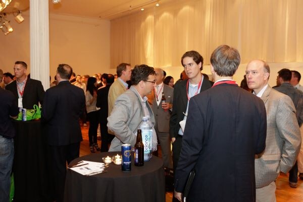 Attendees in suits and jackets stand talking in a crowded room. On a black-clothed table in front are a balance water bottle, a beer bottle, a can and a lit candle.