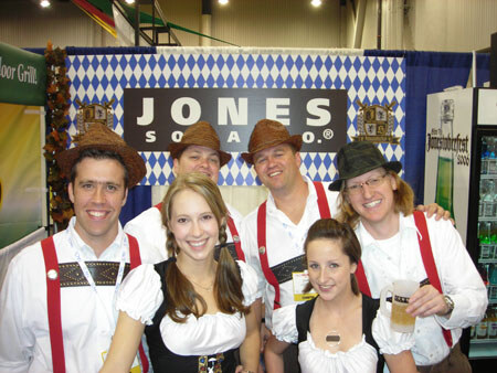 Five people in Oktoberfest-style outfits, including felt hats and red suspenders, pose in front of a Jones Soda sign with a blue and white diamond pattern. One holds a glass of beer.