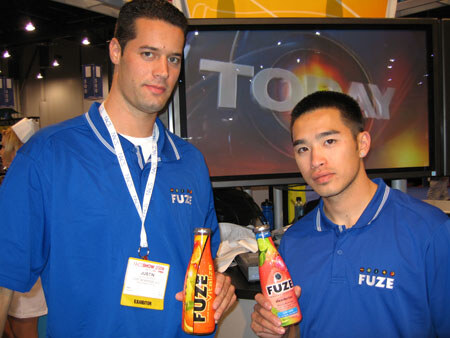 Two men in blue Fuze polo shirts each hold a bottle of Fuze beverage. The man on the left wears an exhibitor badge reading Justin. A TV screen is behind them.