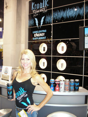 A smiling blonde woman in a black Kronik Energy tank top holds a can at a counter, with a Kronik Energy wall display and several cans on the counter beside her.