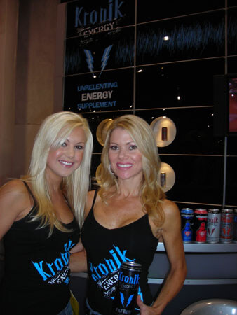Two smiling blonde women in black Kronik Energy tank tops stand together in front of a Kronik Energy wall sign reading Unrelenting Energy Supplement, with cans on a counter.