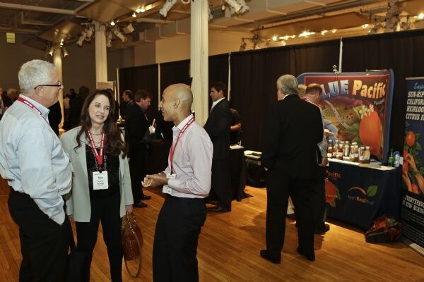 Attendees with red lanyards talk in an exhibit hall in the foreground, while a Blue Pacific booth with a banner and bottles on a table stands in the background.
