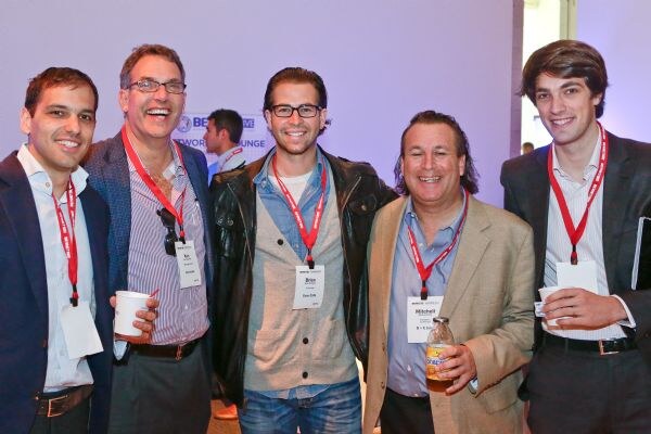 Five smiling men in blazers and jackets stand close together wearing red lanyards and name badges. Brian is in the center in a black leather jacket and glasses; Mitchell is right of center holding a bottle.