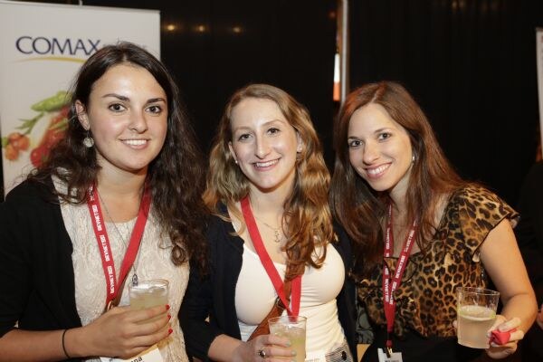 Three smiling women with red lanyards hold glasses of drinks, standing in front of a COMAX banner showing vegetables.