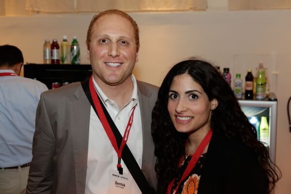 A smiling man wearing a badge reading Derrick and a red lanyard poses beside a dark-haired woman in a red lanyard. Shelves of bottled beverages are visible behind them.