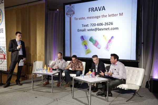 A man with a microphone stands beside three seated men on a stage with bottles on low tables, while a screen shows FRAVA with voting instructions and images of bottles.