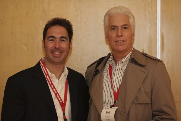Two men stand side by side smiling, both wearing red lanyards. The man on the right, Kevin, wears a tan trench coat over a striped shirt; the man on the left wears a dark blazer.