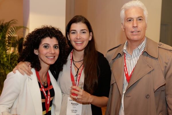 Pamela of Little Big Brands stands in the center with an arm around a curly-haired woman in a white jacket. A gray-haired man in a tan trench coat stands at right; all wear red lanyards.