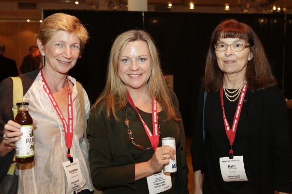 Three women wearing red lanyards stand together smiling. Penelope on the left holds a bottled drink, the woman in the middle holds a can, and Patti stands on the right wearing a layered necklace.