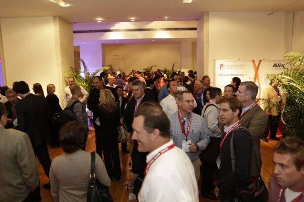 A crowded networking reception of attendees wearing red lanyards stands in a room with plants, and a Pharmachem Laboratories banner is visible on the right wall.